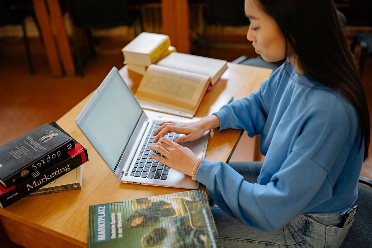 Student typing on a laptop at a wooden desk beside marketing textbooks and open study books
