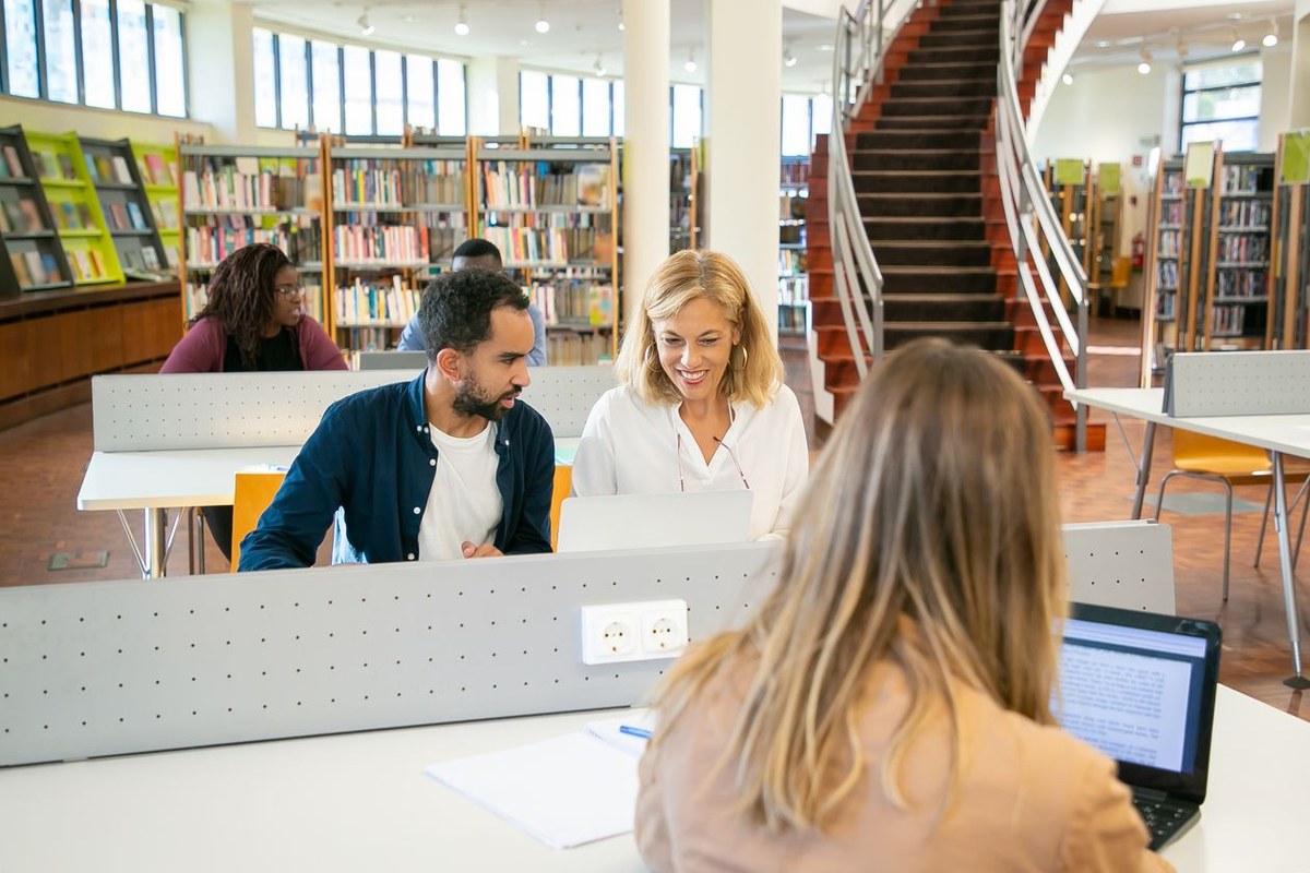 Students collaborating over a laptop at a white desk in a bright multi-level university library
